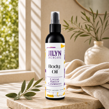 Load image into Gallery viewer, A bottle of JILYN SKINCARE Body Oil in a warm, styled home setting. The black spray bottle with a clear cap stands on a beige stone slab next to a beige linen cloth, green leaves, and a soft-focus background with a window and plant.

