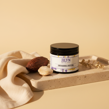 Load image into Gallery viewer, Jilyn Skincare CocoaShea Butter 2oz black jar lifestyle photo on stone tray with cocoa and shea props
